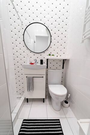 Part of a small new modern bathroom. The main colors - white and black. Tiled walls and floors. Cabinet with sink, shelf and mirror on the wall, toilet, toilet tank, a rug on the floorの写真素材