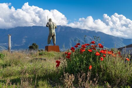 View from the ruins of the city of Pompeii to the mountains and clouds on a clear evening in spring, Italy. In the center is a sculpture Daedalus by sculptor Igor Mitoraj.Red poppies bloom next to itの写真素材