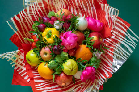 A vibrant multicolored bouquet of vegetables and fruits with pink peonies and green leaves wrapped in corrugated red paper and striped clear cellophane on a lush green.の写真素材