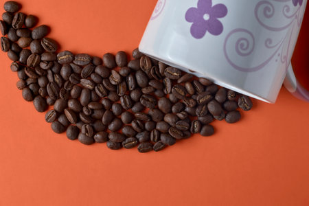 Roasted coffee beans pouring out of a coffee cup with an orange backgroundの写真素材