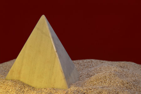 A smooth marble pyramid sculpture partially buried in sand with a dark background, evoking themes of tranquility and structure.の写真素材