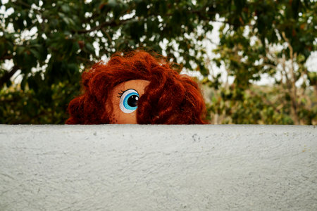 A playful depiction of a doll with curly red hair and bright eyes peeking over a wall, set against a...