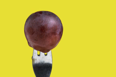 A striking close-up photograph of a vibrant red grape pierced by a metal fork against a yellow backgroundの写真素材