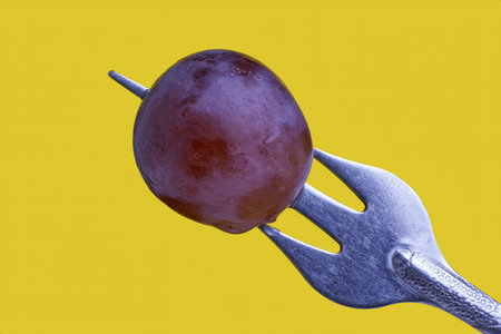 Close-up of a vibrant red grape pierced by a metal fork on a yellow backgroundの写真素材