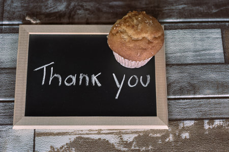 A golden muffin rests on a chalkboard that reads 'Thank you', set on a rustic wooden table.の写真素材