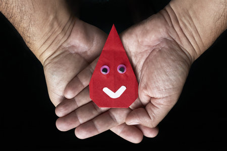 An origami drop of blood with bulging eyes and a white smile remains in open hands, evoking the importance of blood donation.の写真素材