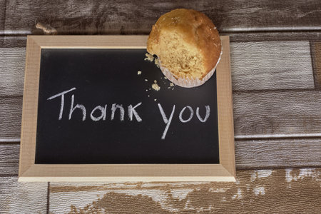 A bitten muffin remains on a chalkboard reading "Thank You," placed on a rustic wooden table.の写真素材