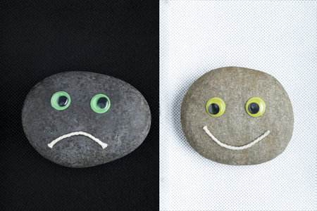 Two smooth stones with bright googly eyes show opposite moods, sad on a dark background and happy on a light background.の写真素材