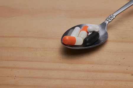 Assorted capsules and tablets sit in a vintage silver spoon on a wooden surface, suggesting medication, health, and daily dosage. Ideal for wellness, pharmacy, and medical product visuals.の写真素材