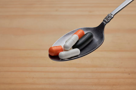 Close-up of assorted capsules and pills on a silver spoon over a wooden backdrop, conveying medication, healthcare, and wellness concepts with a neutral, everyday clinical feel.の写真素材
