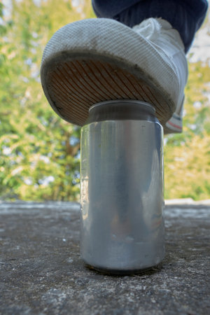 A sneaker presses down on a silver aluminum can resting on a concrete surface, with a blurred green park backdrop. The scene conveys action, balance, and outdoor exploration.の写真素材
