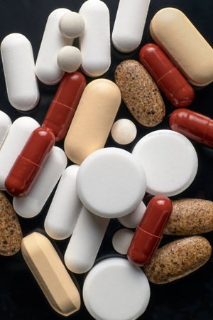 Close-up of white tablets, red capsules, beige caplets and speckled pills scattered on a dark surface.の写真素材