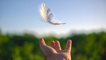 A delicate white feather gently descends towards an outstretched hand, set against a backdrop of lush greenery and a clear blue sky, evoking a sense of peace and lightness.の素材
