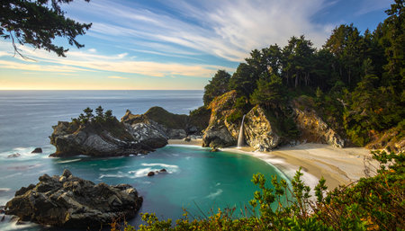 A picturesque view of a secluded beach where a waterfall meets the Pacific Ocean. Lush greenery covers the cliffs surrounding the pristine turquoise water.の素材