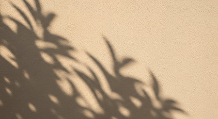Abstract background featuring the soft, blurred shadow of plant foliage cast by the sun onto a warm, neutral beige wall creating a calm and serene atmosphere.の素材