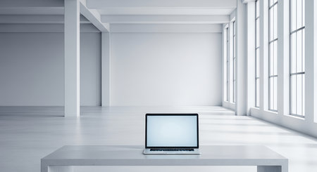 A laptop with a blank white screen sits on a table in a vast, empty, and bright industrial loft. The minimalist interior features white walls, floors, and columns.の素材