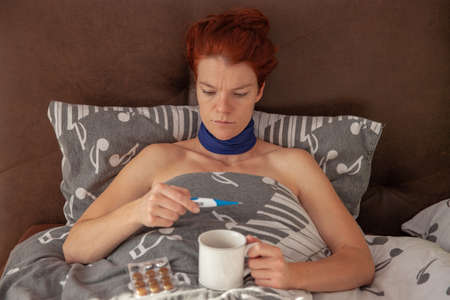 Young sick woman in bed checks the temperature on the thermometer. Hot tea and pills to combat the...