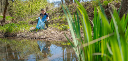 collecting garbage in a plastic bag, a volunteer man helps in the forest on the river bank.