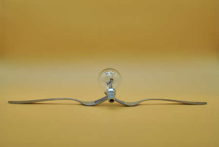 light bulb balancing on a composition of 2 intertwined forks isolated on orange neutral background, conceptual photo to indicate the right division of electricityの写真素材