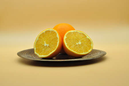 An orange cut in half and a whole orange placed on an elegant and antique silver plate on orange neutral backgroundの写真素材