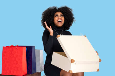 YOUNG WOMAN SMILING HAPPILY AS SHE OPENS GIFT BOX, SITTING DOWN AND BESIDE HER SEVERAL SHOPPING BAGS