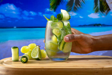 Refreshing cocktail glass decorated with grapes, cucumber and lemon, held by a male hand on a beach backgroundの写真素材