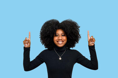 Smiling positive young woman with afro hairdo, indicates with both index fingers, wearing casual black dress, feeling satisfied, isolated on blue background People and promotion conceptの写真素材