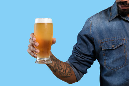 cropped shot of tattooed man holding glass of beer isolated on blueの写真素材