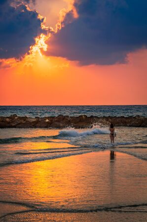 A young woman standing between the waves and admiring the sunset over the sea.のeditorial素材