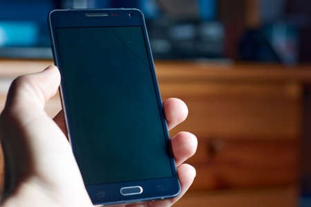 Hand holding broken mobile phone with small crack on black screen which prevents it from working.