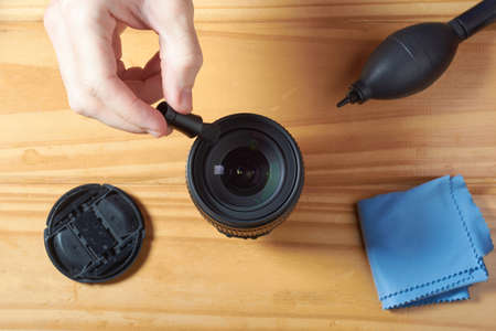 Top view of male hand using small cleaning brush to remove dust from DSLR camera lens with air blower, cleaning cloth and lens cap on wooden table.の写真素材