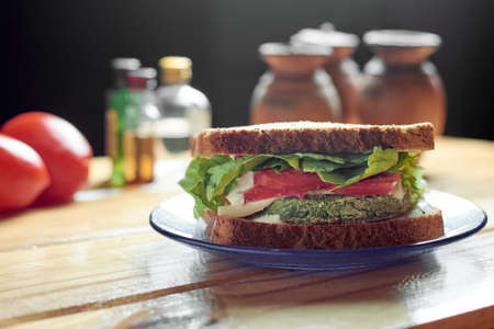 Vegan sandwich with medallion of spinach and carrot, tomato, lettuce, vegan cheese and mayonnaise on rustic wooden table, surrounded by tomatoes, spices and condiments. Bread with poppy, sunflower and flax seeds, and oat.の写真素材