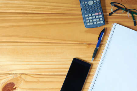 Top view of workplace with mobile phone, pen, blank notebook, calculator and glasses on rustic...