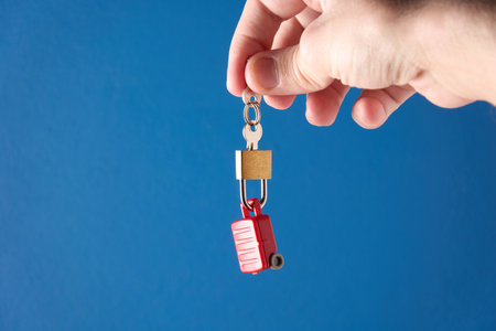 Male hand holding keys and padlock locked on miniature toy suitcase. Concept of travel insurance, safe travel. Blue background with copy space.の写真素材