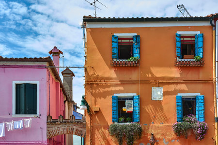 Horizontal view of picturesque facades of buildings with vibrant orange, blue and pink colors under blue sky with clouds on sunny spring day in Burano, Italy.のeditorial素材
