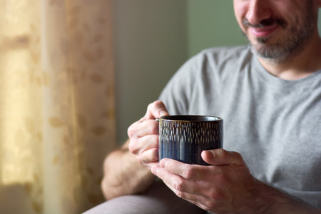 Close up of young caucasian man smiling and holding a cup of tea. Joyful man enjoying hot tea in his...