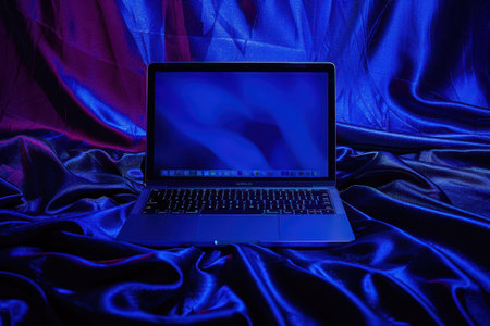 Laptop glowing on blue satin fabric with blank screen. AI-generated.の素材