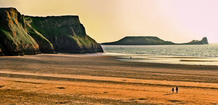 View of the beach in Rhossili Bay, Wales, during sunset, with couple walking in the distance and...