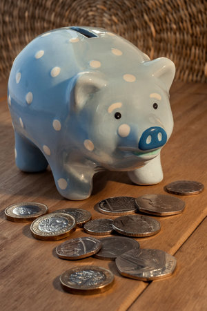 Light blue piggy bank with white dots, and British sterling pound coins around it on a light wooden surface. Money saving concept. home financeの写真素材