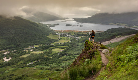 Woman standing on a rock with hiking poles overlooking the valley of Glencoe, Highlands of Scotland, with Loch Leven and village in background and atmospheric low hanging cloudsの写真素材
