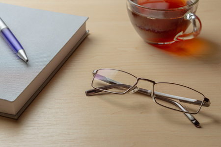 Eyeglasses, diary, blue pen and cup of tea on a brown table, selective focusの写真素材