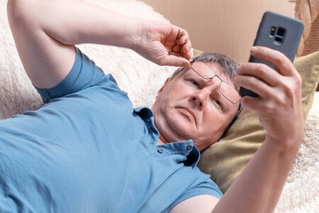 A man with glasses looks at a smartphone while lying in a cozy sofa. Middle-aged businessman checks mail in a smartphoneの写真素材