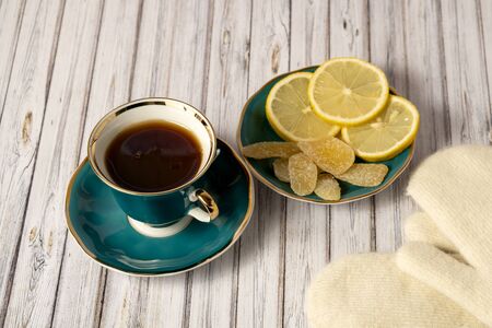 Green cup of hot tea, saucer with lemons and candied ginger and white mittens on a wooden tableの写真素材