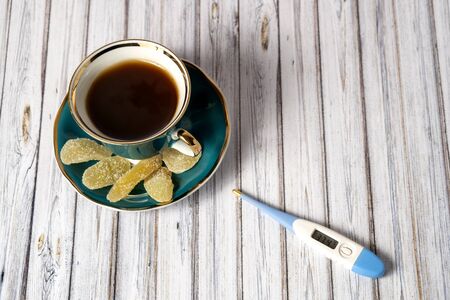 Thermometer, green cup of hot tea and saucer with lemons and candied ginger on a wooden table. Top...