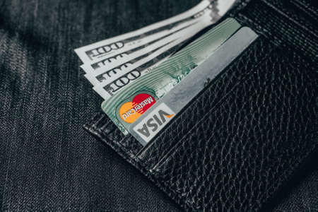 Odessa, Ukraine - April 12, 2020: Black genuine leather wallet with banknotes and credit card inside, jeans background. Soft focusのeditorial素材