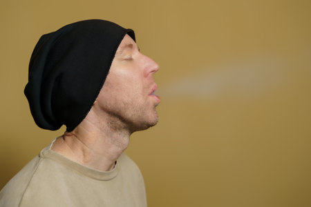 A man with closed eyes in a black hat releases a cloud of smoke. Close-up portrait of a white man in a black cap.の写真素材