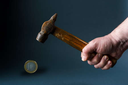 A man hits a one euro coin with an old rusty hammer. Male hand with hammer and euro coin close up on gray background.の写真素材