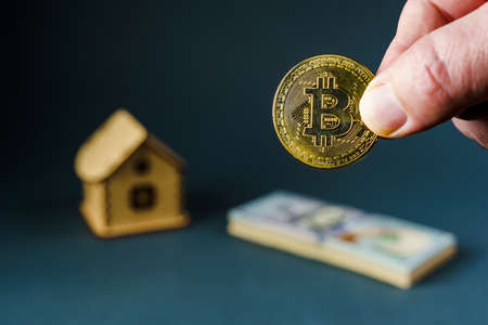 Bitcoin coin in hand against the background of a toy wooden house and a stack of one hundred dollar bills, soft focusの写真素材