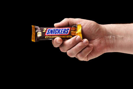Hand holding a chocolate bar close-up. A man offers a Snickers chocolate bar, black backgroundのeditorial素材