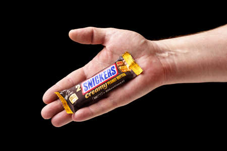 A man offers a Snickers chocolate bar, hand holding a chocolate bar close-up, black background.